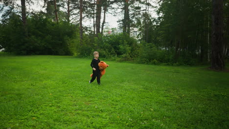young boy dressed in black walking across vast grassy field while holding large orange teddy bear with peaceful forest background and calm natural scenery under soft daylight