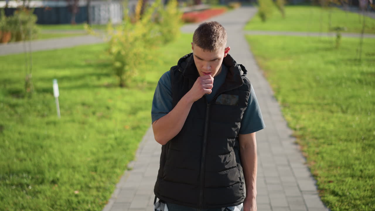 white boy in black vest walks on paved path in green park under bright sunlight inhales vape and coughs while holding chest and looking down at small vape device with discomfort expression