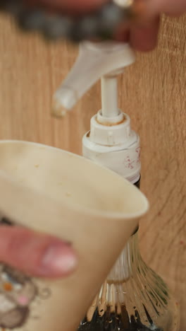 vista de una mano que lleva un brazalete de cuentas y una banda de mano de plata sosteniendo una taza de café mientras bombea jarabe de café. capturando el proceso detallado de agregar jarabe a la taza de cafe
