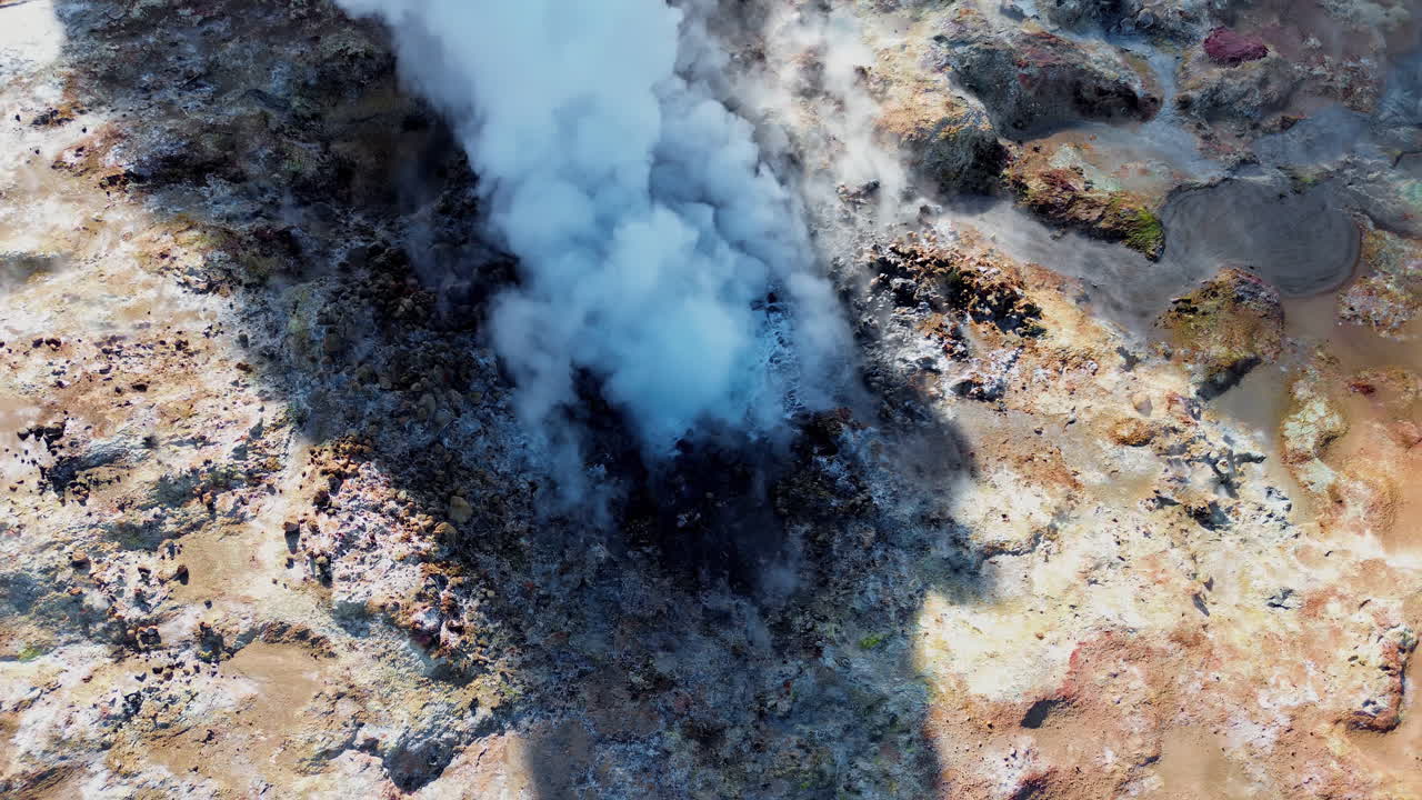 Vista aerea con drone di fumarole geotermiche fumanti e pozze di fango bollente in un campo geotermico vulcanico in Islanda. Il terreno ricco di minerali, nelle sfumature del beige, rosso e blu, circonda fumarole attive che rilasciano vapore.
