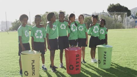 portrait of happy diverse schoolchildren with recycling bins in sports field at elementary school