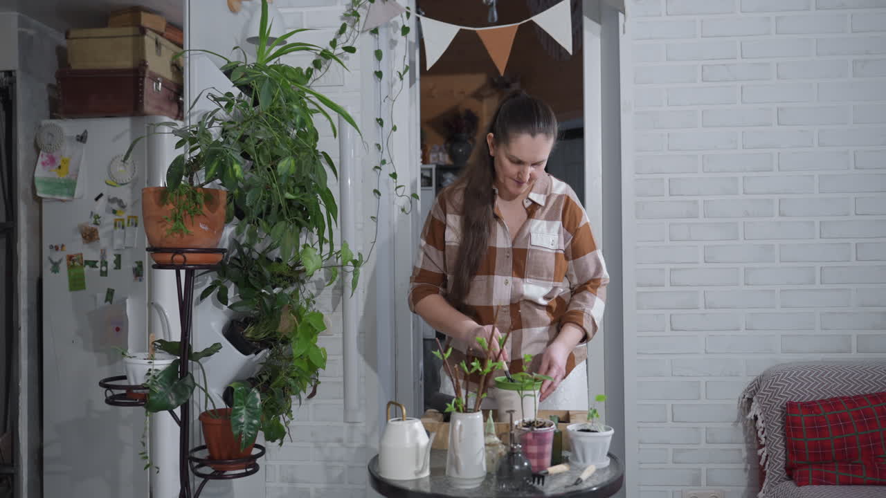 Plant enthusiast using small shovel to dig hole in plastic pot filled with dark soil while gently placing young rooted plant into hole on indoor glass table surrounded by lush potted greenery