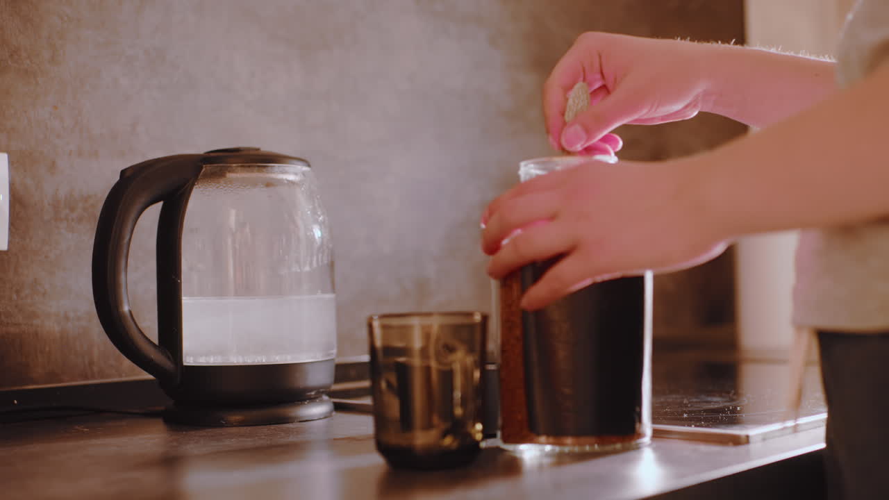 Close up of woman removing lid from coffee container as she prepares to scoop instant granules into cup placed beside electric kettle on counter during early morning beverage routine at home kitchen