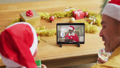 Kaukasischer-Vater-Und-Sohn-Mit-Weihnachtsmützen,-Die-Ein-Tablet-Für-Einen-Weihnachtsvideoanruf-Mit-Dem-Weihnachtsmann-Auf-Dem-Bildschirm-Nutzen