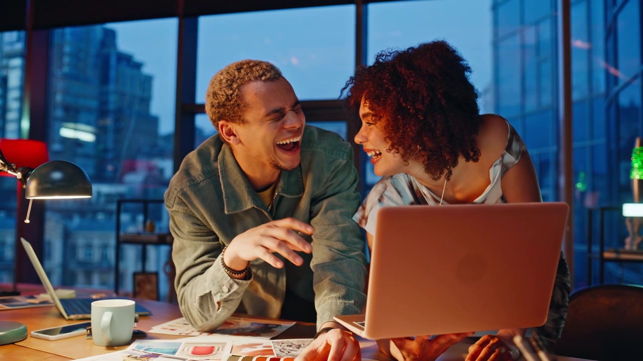 Coworkers laughing over laptop night office. Friends brainstorm design project