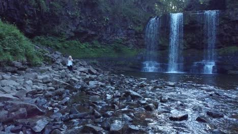 drone flying over river towards a waterfall past a girl