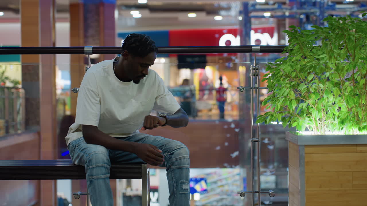 Man in white shirt and jeans seated on bench in shopping mall interior tapping hands together checking time while waiting for someone modern retail lifestyle