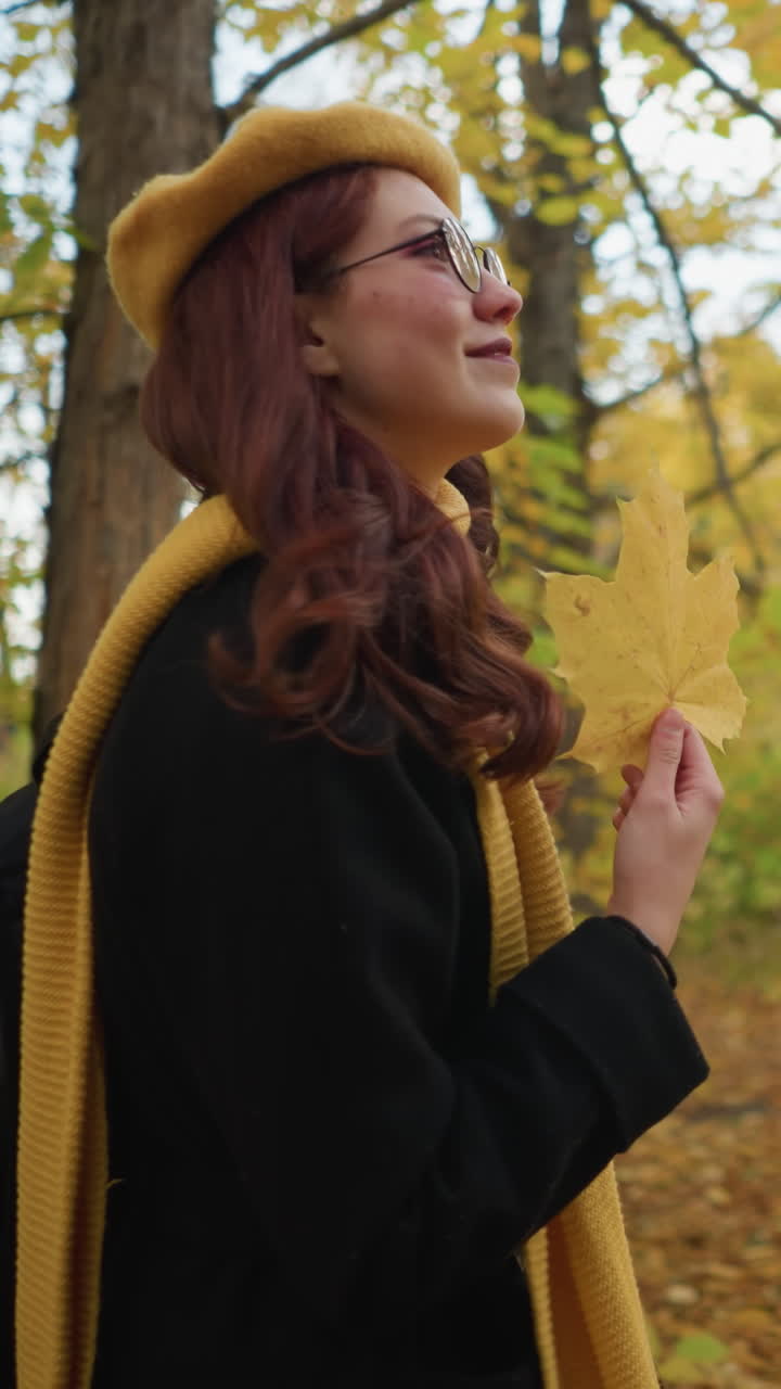 따뜻한 미소를 지으며 가을 잎을 들고 황금 잎자루로 둘러싸인 숲을 걸으며 평화로운 자연과 신선한 가을 공기를 즐기고 있는 여자의 측면 풍경