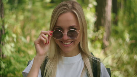 a young female traveler in sunglasses looks directly at the camera and smiles flirting a traveler with a backpack in the park and in the forest in slow motion