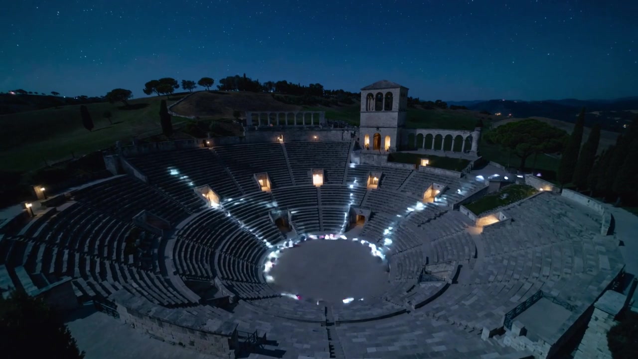 Gruppo che accende le luci, cammina lungo le navate di un antico anfiteatro di notte, formando un anello centrale illuminato