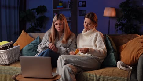 Creative Knitting Evening With Girlfriends on a Cozy Night. Two women enjoying a relaxing evening at home, knitting and laughing on the couch. The friends watch a video lesson on the laptop and learn to knit new patterns with needles