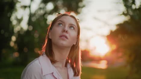 young lady in pink top with glasses resting on her head, warm smile, looks up thoughtfully while bathed in sunlight, sun flares and blurred greenery in background