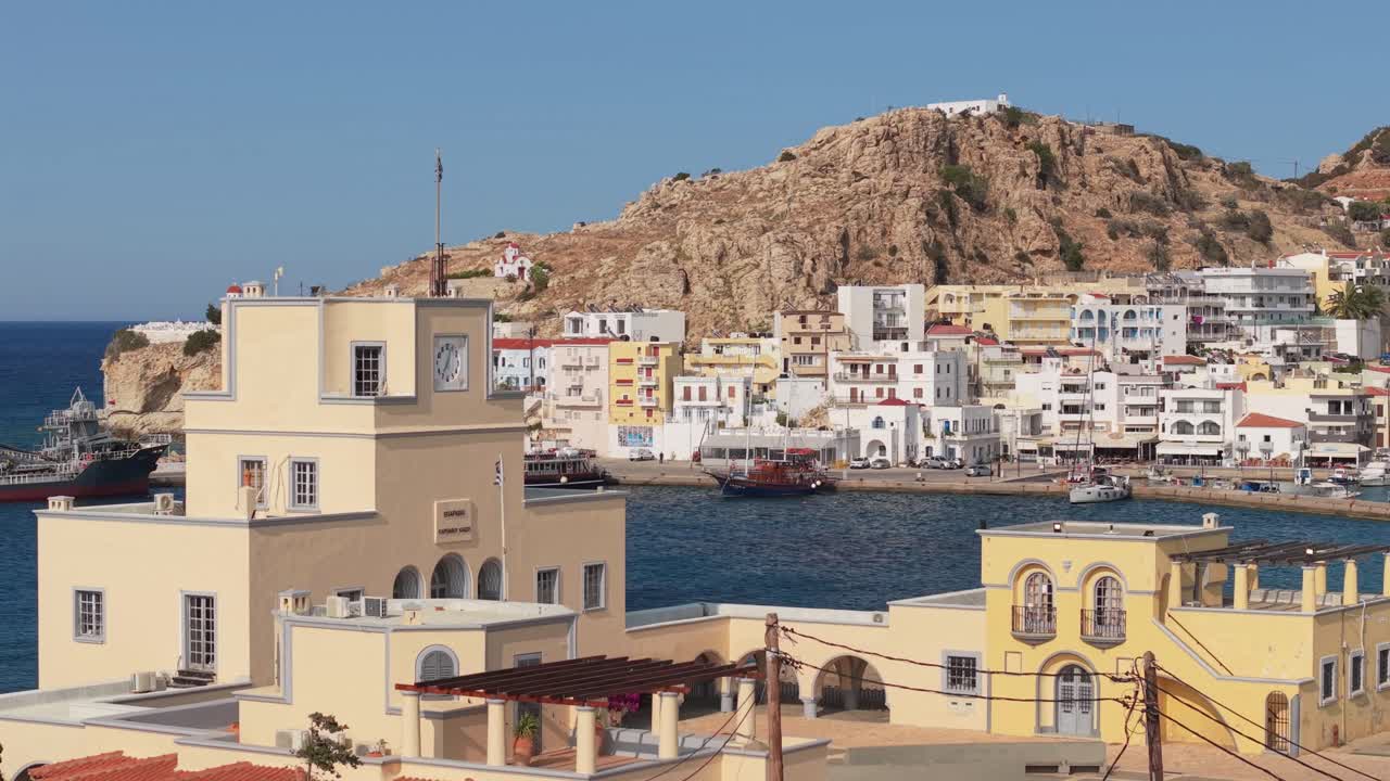 Drone footage revealing the Pigadia seafront behind the Eparchy building, capturing the harbor, waterfront town, and coastal beauty of Karpathos island under a clear sunny sky