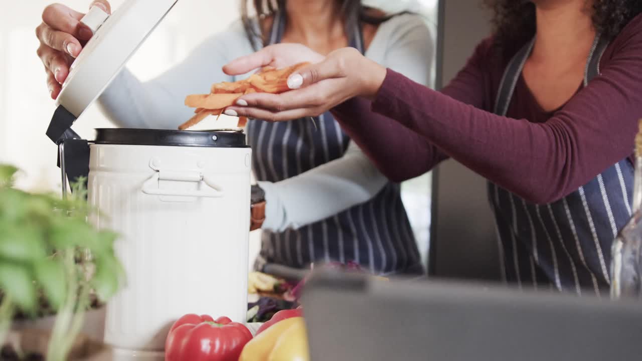 sección media de una pareja lesbiana biracial en delantales compostando residuos vegetales en la cocina, cámara lenta
