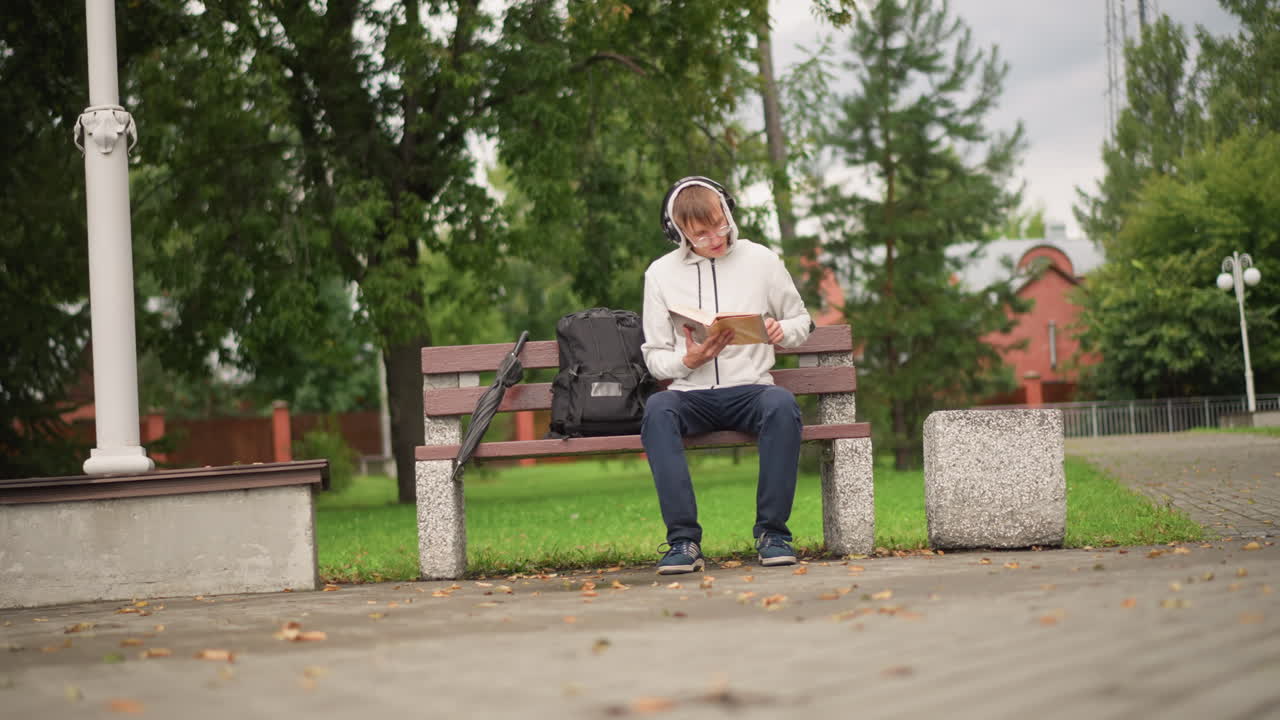 공원 벤치에서 기타를 치는 남성, 흐리고 비 오는 오후, 헤드폰과 후드티 착용, 옆에는 백팩, 조약돌 길에는 낙엽, 고요하고 사색적인 분위기, 은은한 거리 음악, 솔로