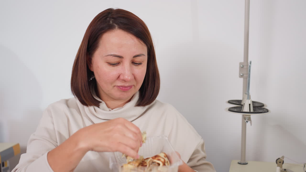 Female tailor seated beside sewing machine in bright organized tailoring workspace eating pancake from plate, enjoying light snack during break time surrounded by fabrics, and sewing equipment