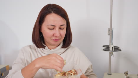 female tailor seated beside sewing machine in bright organized tailoring workspace eating pancake from plate, enjoying light snack during break time surrounded by fabrics, and sewing equipment