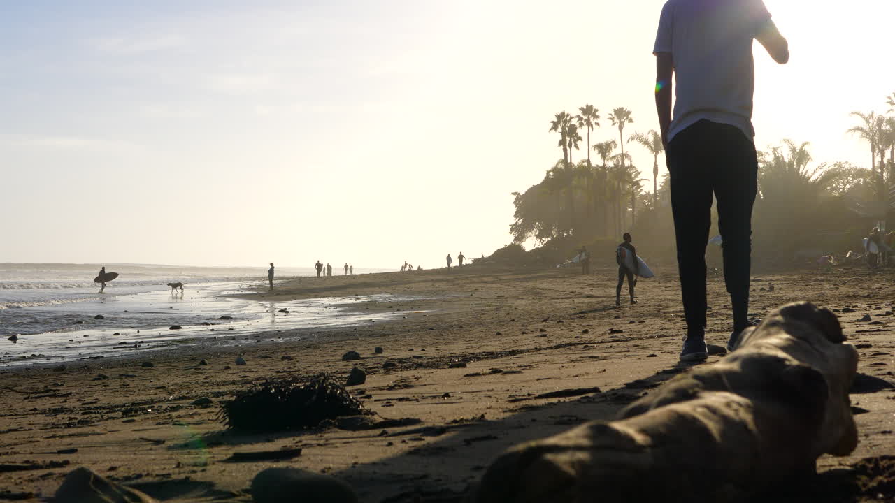 Um homem atraente caminhando na praia no ponto Rincon, na Califórnia, ao pôr do sol, com pessoas e palmeiras em silhueta na hora dourada.