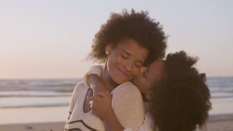 cute girl hugging her mother from behind and kissing her cheek gently while sitting on the beach at sunset