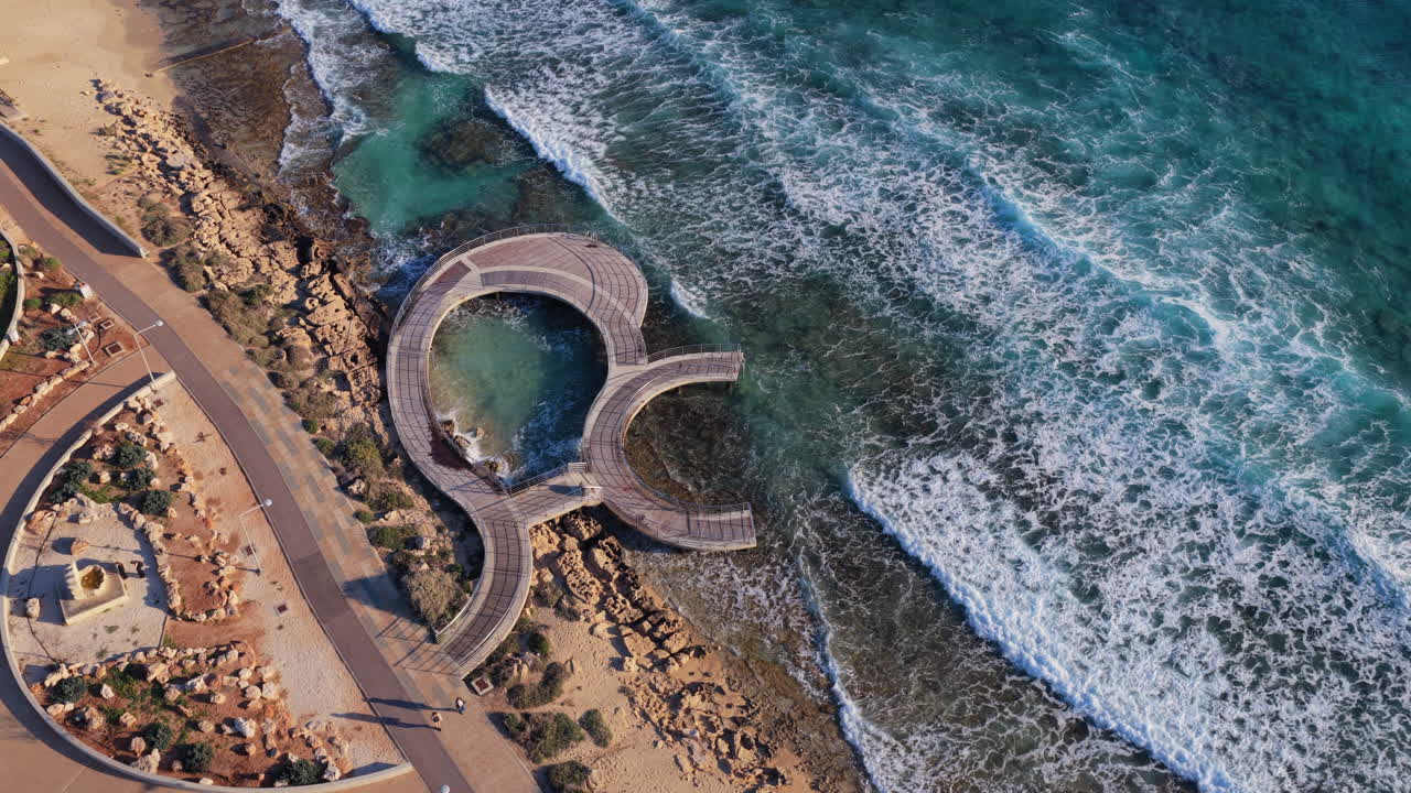 Veduta aerea con drone di una passerella costiera e una piattaforma panoramica curva lungo il litorale ad Ayia Napa, Cipro, con le onde che si infrangono sugli scogli.