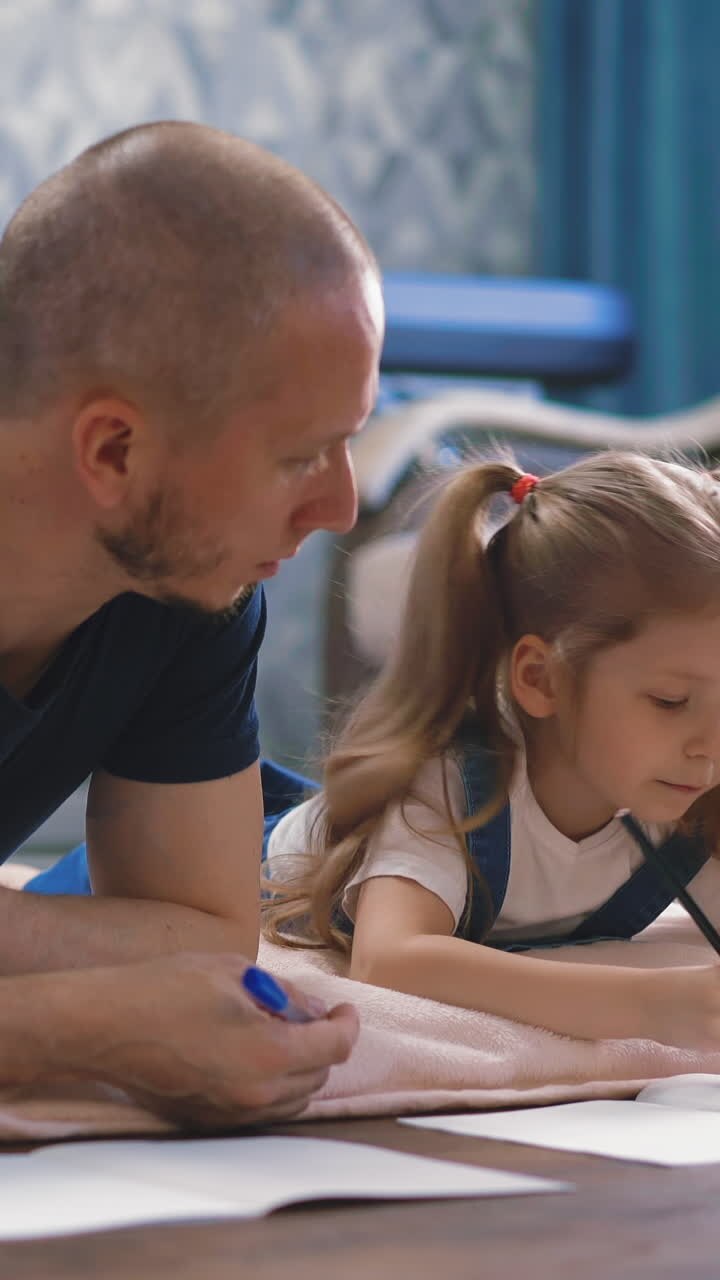 concept of distance learning: young father and student daughter lie on floor in living room on plaid engaged in home schooling, draws in notebook, prompts, help