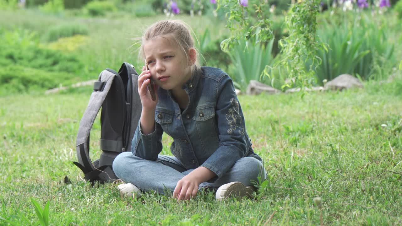 학교에 다니는 어린 소녀가 공원 잔디에 앉아서 전화로 대화를 나누고 있다.