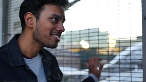 Guy-in-jean-jacket-holding-onto-the-metal-fence-at-a-train-station-has-a-conversation-with-someone-out-of-frame