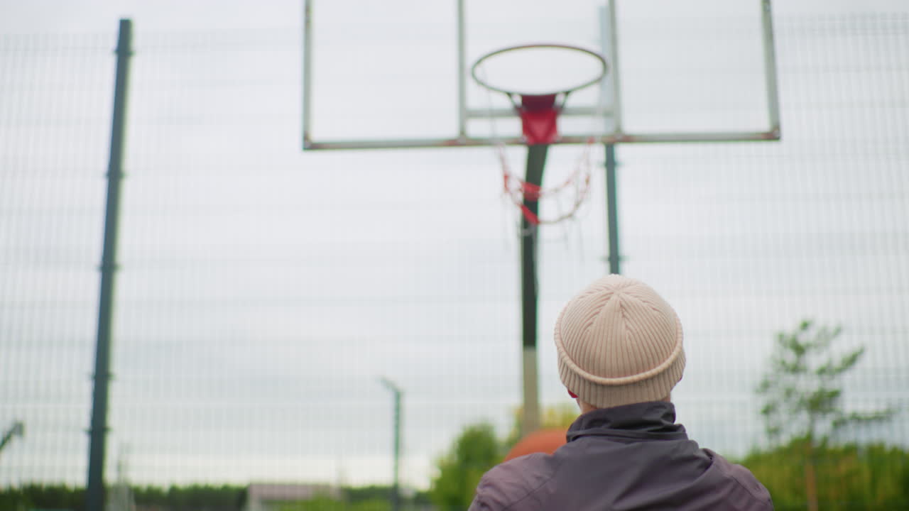 uomo che tira una palla da basket verso il canestro da dietro, prospettiva posteriore ravvicinata con cappello di lana visibile, mani che rilasciano la palla, arco ben definito verso il cerchio e la rete, tabellone e recinzione che inquadrano una scena nuvolosa del parco