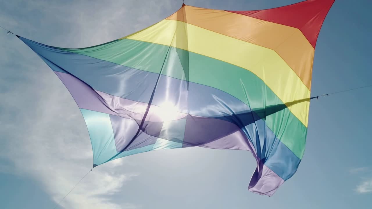 Aquilone triangolare arcobaleno che si gonfia e ondeggia, con le aste che si flettono per il vento nel cielo blu e un bagliore solare in movimento.