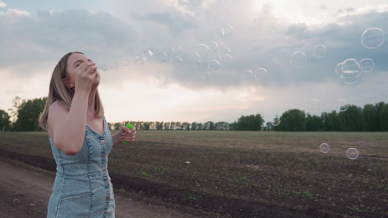 side view of young woman in denim dress walking along dirt road through expansive farmland while blowing iridescent soap bubbles into pastel sky at golden hour