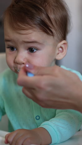 cute baby playing with a metal spoon is fed by mom. messy kid in blue shirt sits at white feeding table eating his food from spoon. blurred backdrop. vertical video