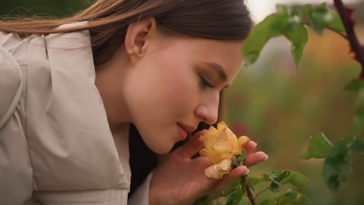 Joven oliendo una rosa amarilla, primer plano del perfil en un jardín otoñal, abrigo color beige, luz dorada suave, rocío en los pétalos, expresión contemplativa, bokeh cinematográfico, ambiente tranquilo, estilo de vida en la naturaleza