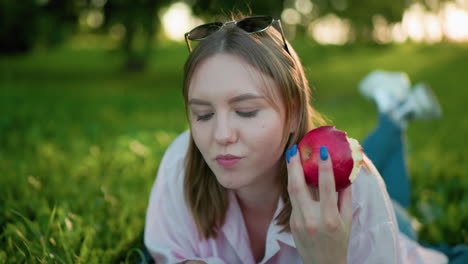 primer plano de una joven comiendo una manzana mientras está acostada en un campo cubierto de hierba, trabajando en una computadora portátil, disfrutando de un momento pacífico con la luz del sol filtrándose a través de los árboles