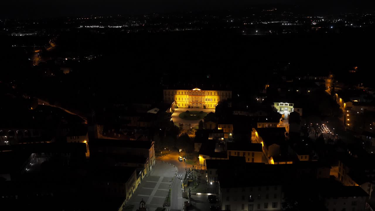 Aerial night view of the illuminated Villa Gallarati Scotti and the surrounding cityscape in Vimercate, Italy. barrel roll shot