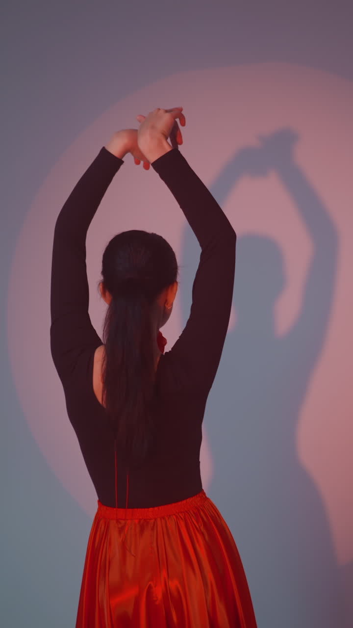 una dama con un vestido negro y rojo se para frente a la pared, moviendo su cuerpo y manos lentamente de una manera de baile contra un fondo gris