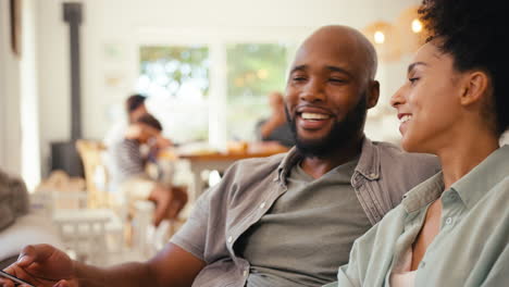 Couple-At-Home-On-Sofa-Streaming-Show-Or-Movie-To-TV-With-Multi-Generation-Family-In-Background