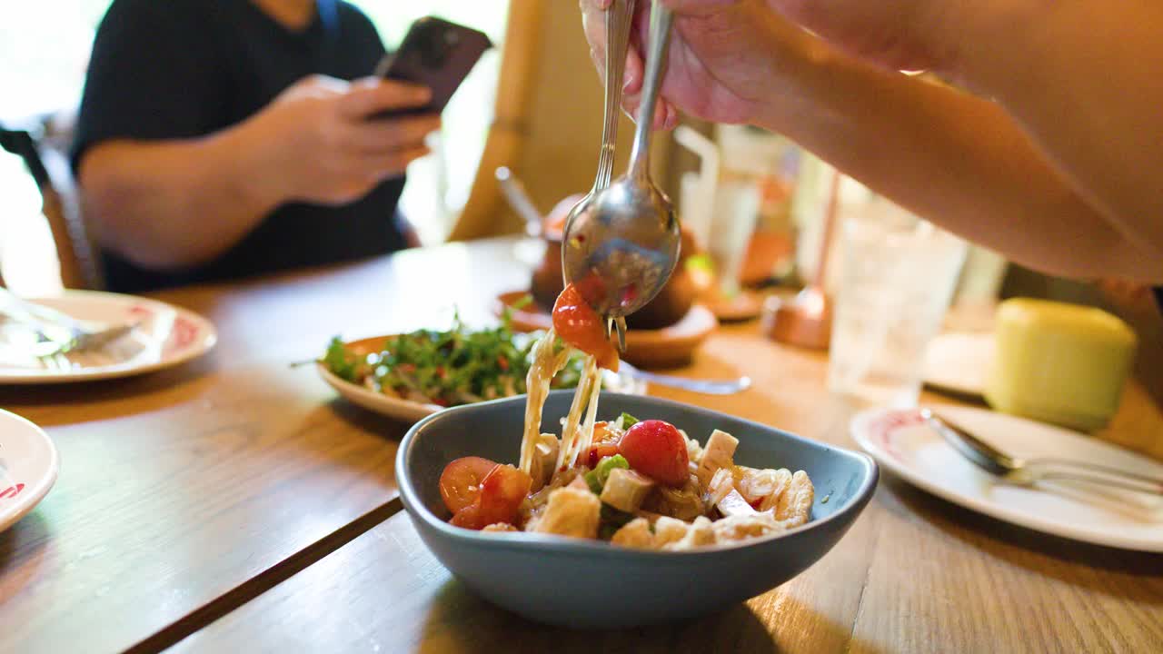 Hand mixes spicy Thai rice vermicelli salad in natural light, casual restaurant dining setting, close-up