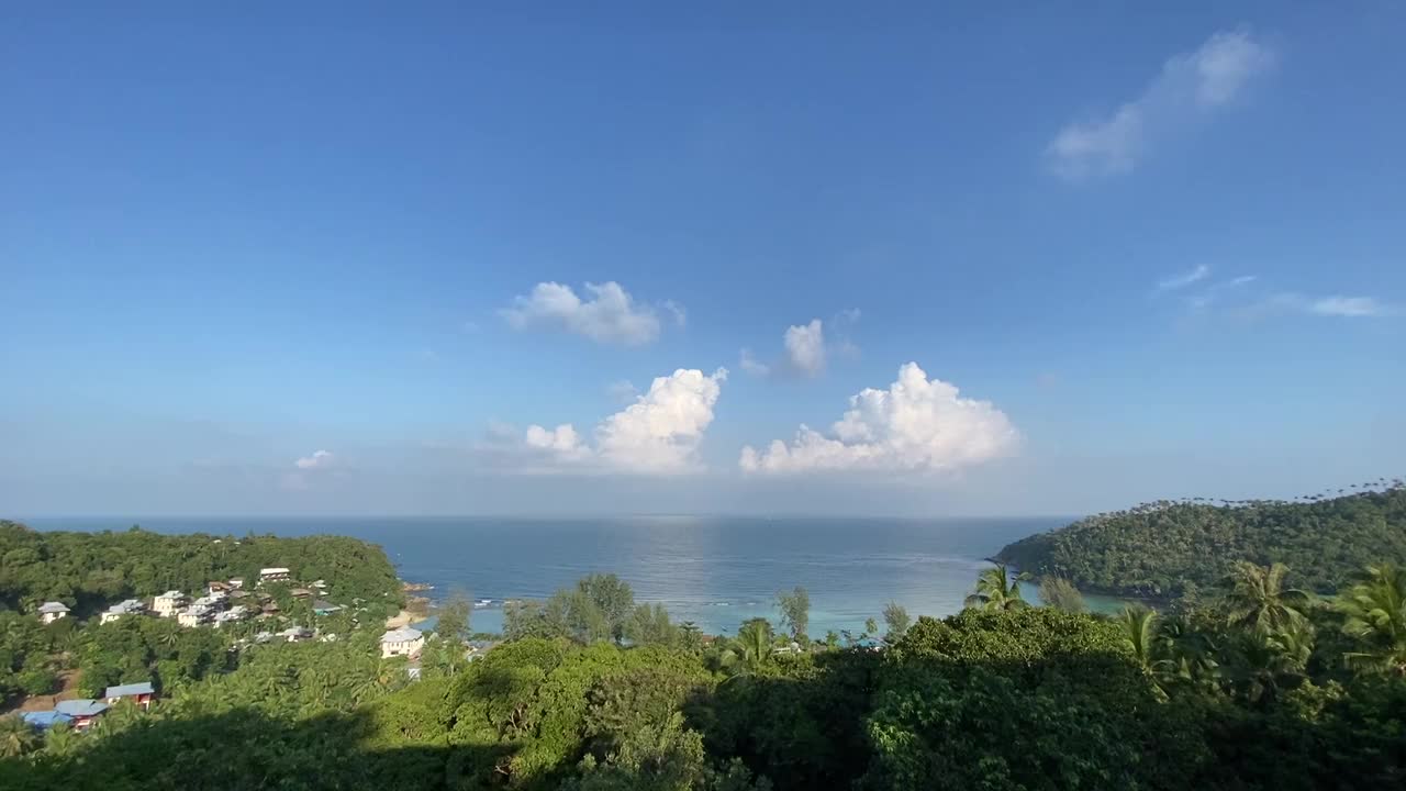 Scenic view of Ao Nang coastline in Krabi, Thailand with turquoise waters, green hills, village rooftops, and bright blue sky dotted with clouds creating a peaceful tropical setting