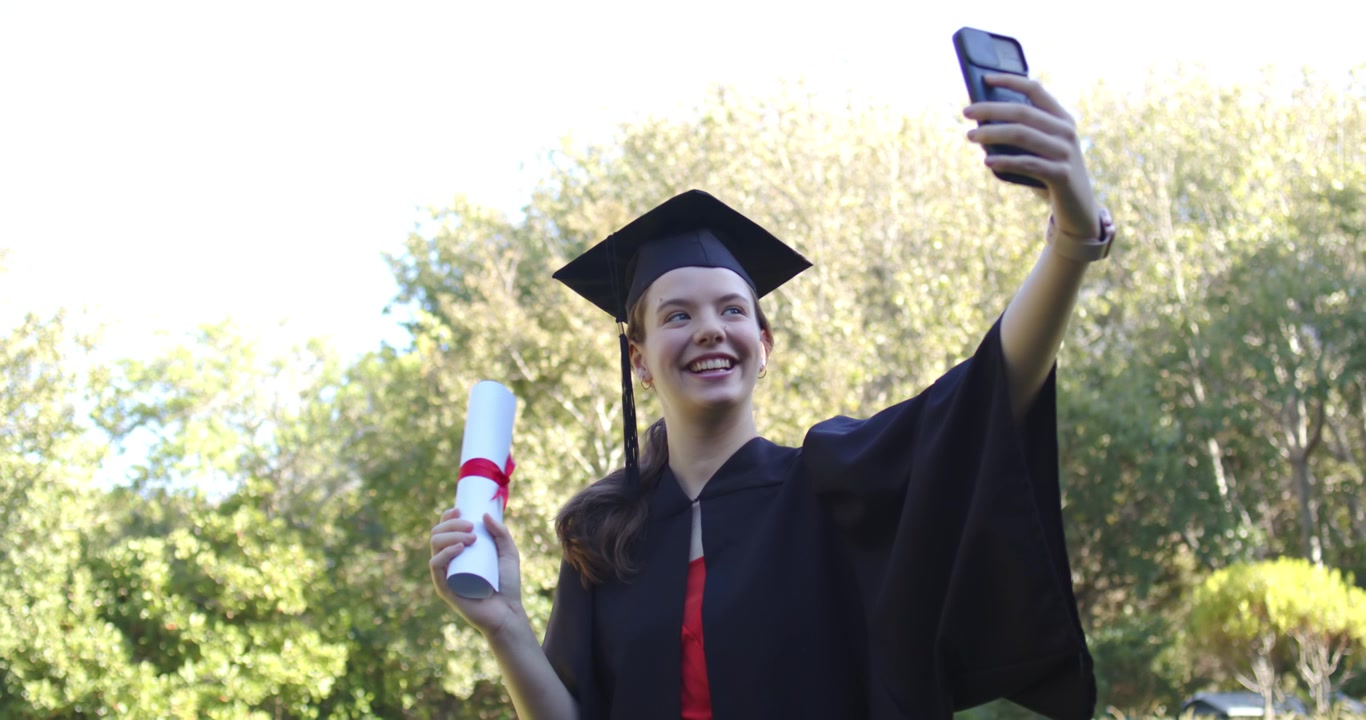 Donna in toga e tocco che si fa un selfie con il telefono al parco dopo la laurea, sventolando il diploma con il nastro.