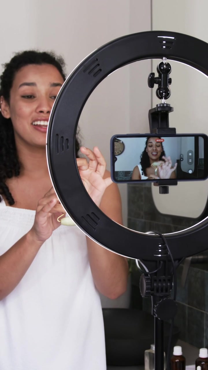 Vertical video: Showing woman rotating roller at vanity, phone in ring light recording in white top