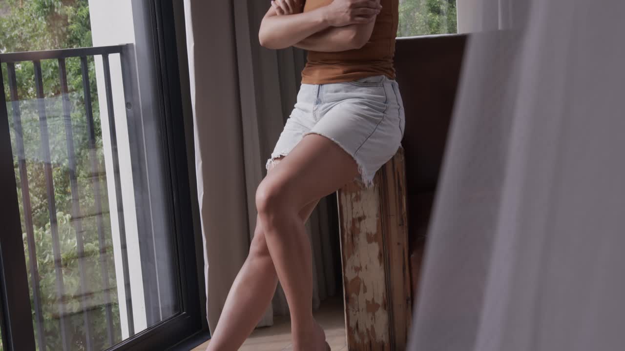La telecamera si allontana mentre una donna si sposta, appoggiata a uno sgabello e guarda oltre il fogliame del balcone.
