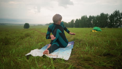 woman in green black suit seated on yoga mat in seated twist pose practicing yoga, in a vast grassy field with trees lined up in the distance under a cloudy sky