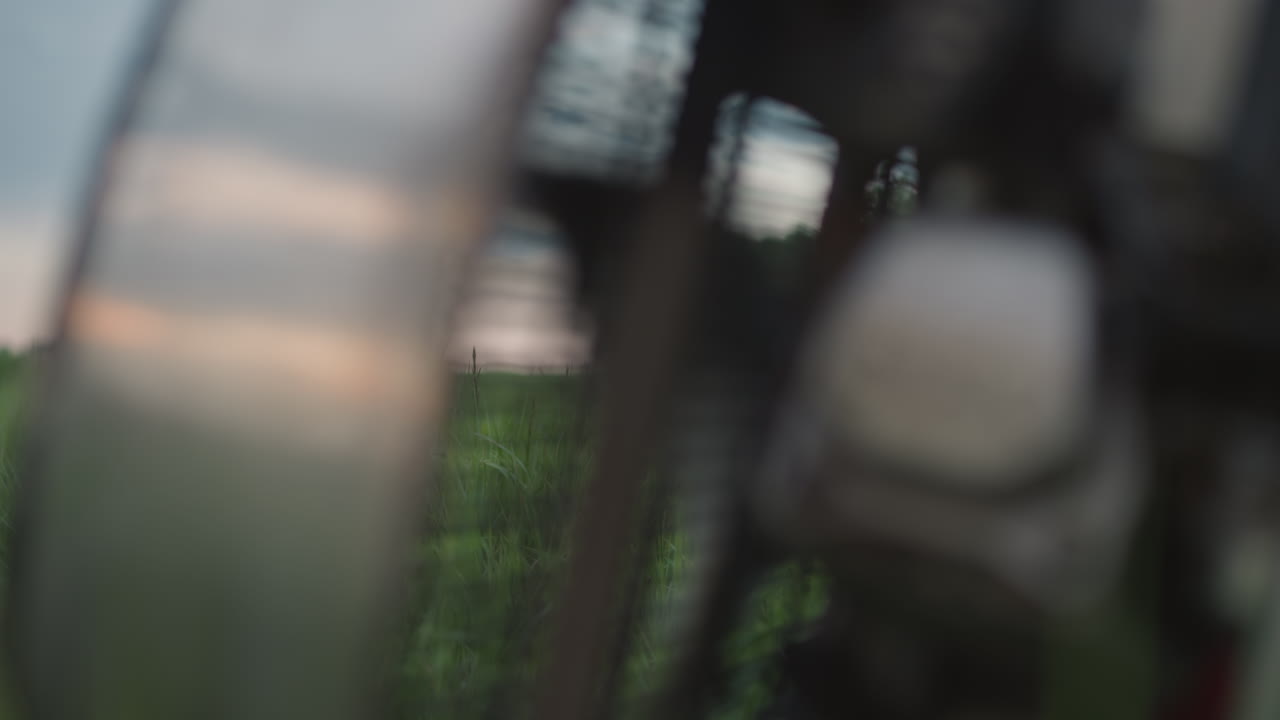 closeup rotating metal fan cage whipping dynamic gusts through tall green grass causing delicate blades to sway gently against blurred countryside backdrop under soft pastel evening sky