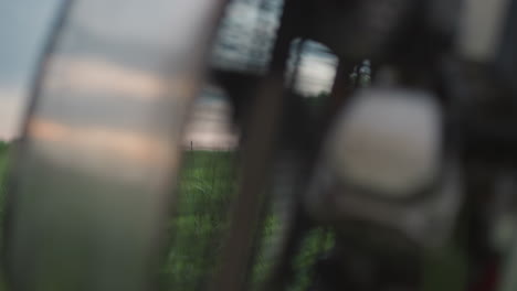 closeup rotating metal fan cage whipping dynamic gusts through tall green grass causing delicate blades to sway gently against blurred countryside backdrop under soft pastel evening sky