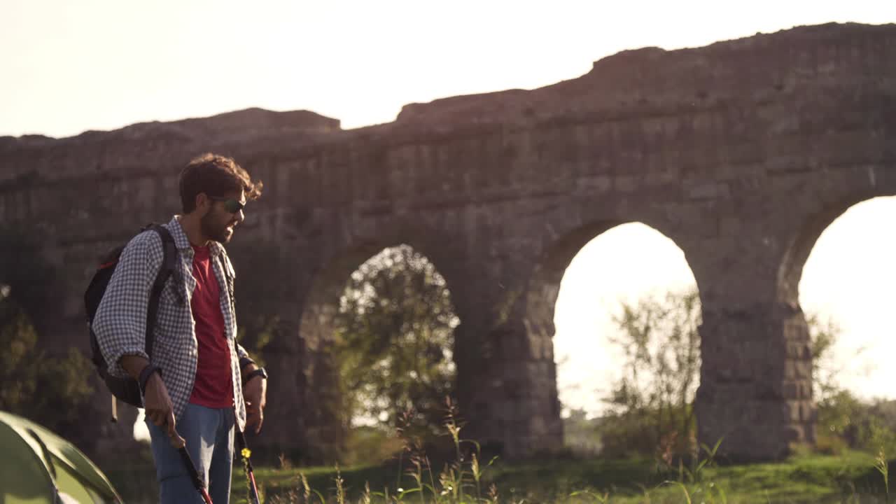 젊은 배 타는 남자가 황혼이 지는 로마의 파르코 델리 아키도티 공원 유적에 있는 로마 수로 앞에서 잔디 위에 지이를 들고 맨발로 고 있다.