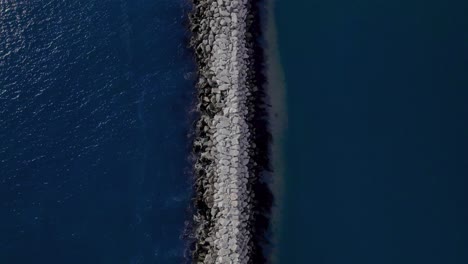 Birdseye-view-following-jetty-separating-blue-ocean-water-and-sand-bank