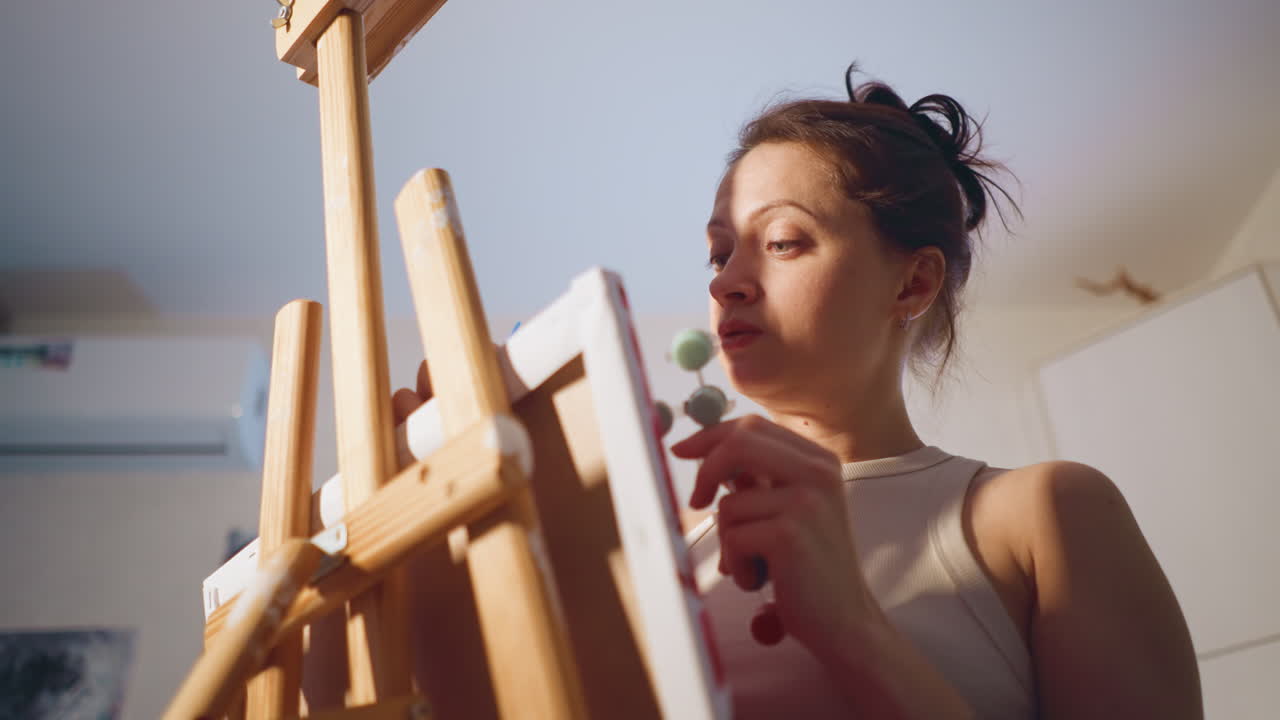 Low Angle White Woman Painting At Easel In Morning Light, Serene Concentration, Soft Shadows, Holding Brush And Palette, Home Studio Backdrop, Thoughtful Expression, Focused Creative Routine