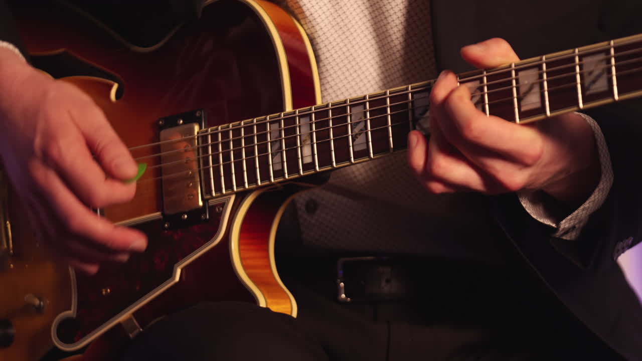 Fixed close-up of a jazz guitarist’s hands playing with a pick, showing strings and guitar body against a blue backdrop