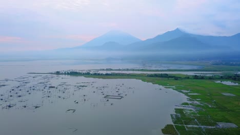 Aerial-view-of-huge-lake-with-fish-cages-in-misty-morning-and-mountain-on-the-background---Rawa-Pening-Lake,-Indonesia