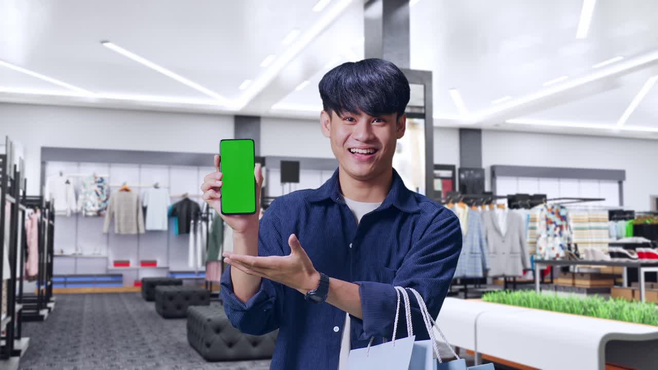 녹색 화면 휴대 전화 포즈를 들고 옷 가게에 서 있는 동안 카메라에 미소 짓는 쇼핑 가방을 들고 있는 아시아 쇼핑 남자